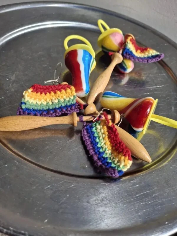 Små hæklede regnbueflag på træpinde ligger sammen med farverige pyntegenstande på en metalbakke.