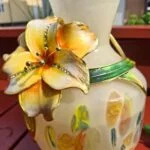 Nærbillede af vasens hals og påsatte gule blomst med grønne blade.