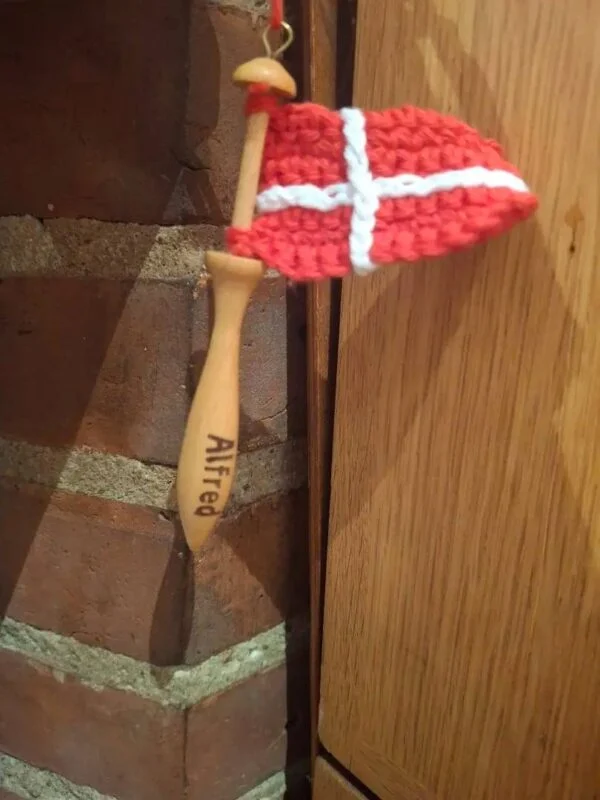 Rødt og hvidt hæklet Dannebrog på en trækniplepind med navnet Alfred, fotograferet ved en murstensvæg.