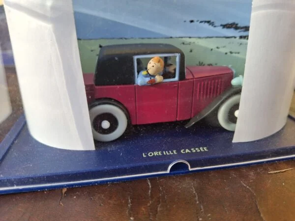 Rød Tintin-modelbil i klar udstillingskappe med illustreret baggrund