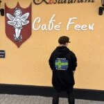 Original sort Kiksede Knud hoodie med Café Feens flagmotiv på ryggen, vist ved caféens gule facade