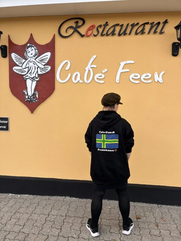 Original sort Kiksede Knud hoodie med Café Feens flagmotiv på ryggen, vist ved caféens gule facade